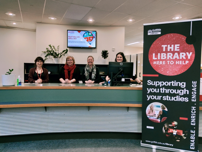 Four smiling library staff stand behind the front desk of South Bank’s Queensland Conservatorium Library. A vertical banner stands beside the desk and reads ‘The Library: Here to help’ and ‘Supporting you through your studies’.