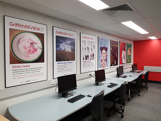 A row of Griffith Review posters on the wall at QCA Library above a row of computers.