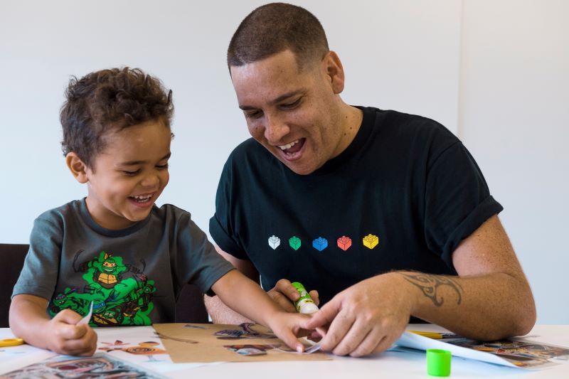 An Indigenous man creating artwork with an Indigenous child. Both people are smiling and having fun.