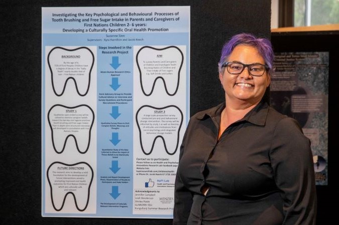 An Indigenous person with short purple hair standing in front of a research poster on ‘Developing a Culturally Specific Oral Health Promotion’.