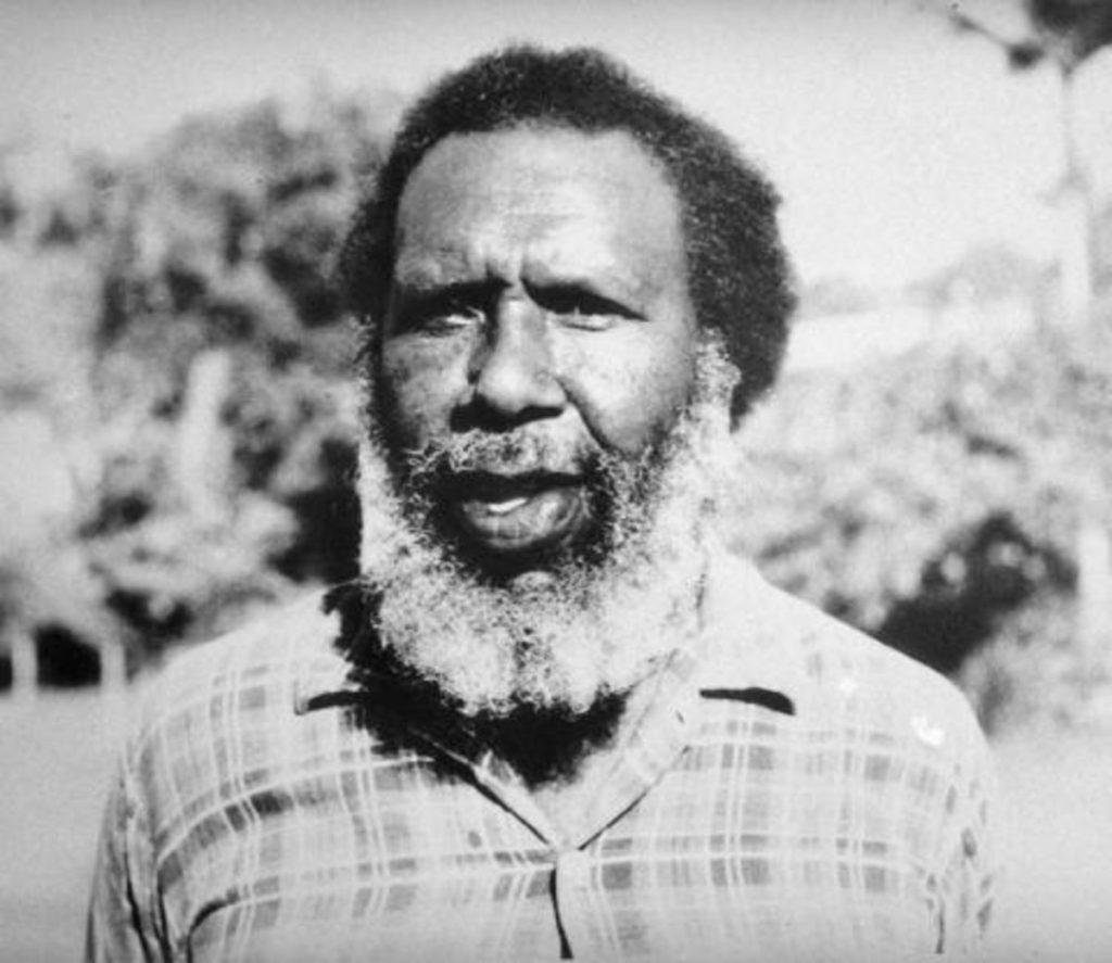 Black and white photo of Eddie Mabo wearing a checkered collared shirt. Trees can be seen in the background.