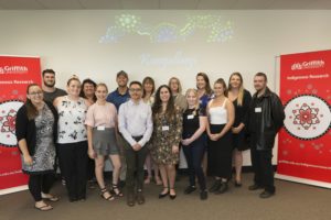 A group of Kungullanji Research Pathways Program participants standing together in front of a projected background and signs to the side that say ‘Griffith Indigenous Research’ 
