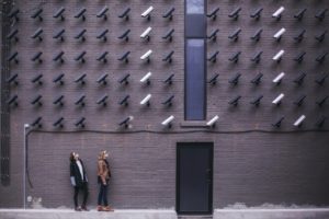 Two people are on a sidewalk staring up at a wall full of security cameras.