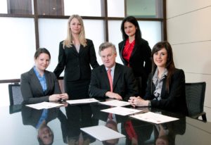 A group of four women and one man are seated and standing around a large conference table. There are papers on the table in front of them.