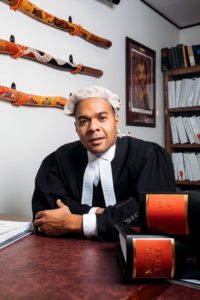 A picture of Joshua Creamer. He is dressed in a barrister's robe and wig is leaning on a desk. There are law books in front of him and three didgeridoos mounted on the wall in the background.