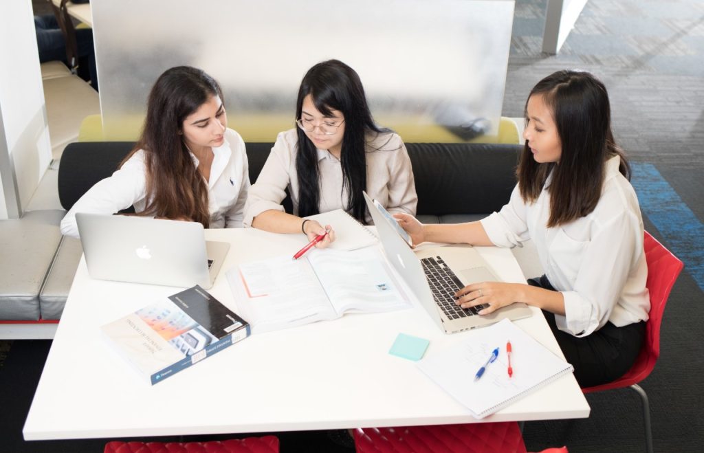 A group of three people are sitting together around a table and talking. There are open laptops, books, notepads and pens on the table in front of them. 