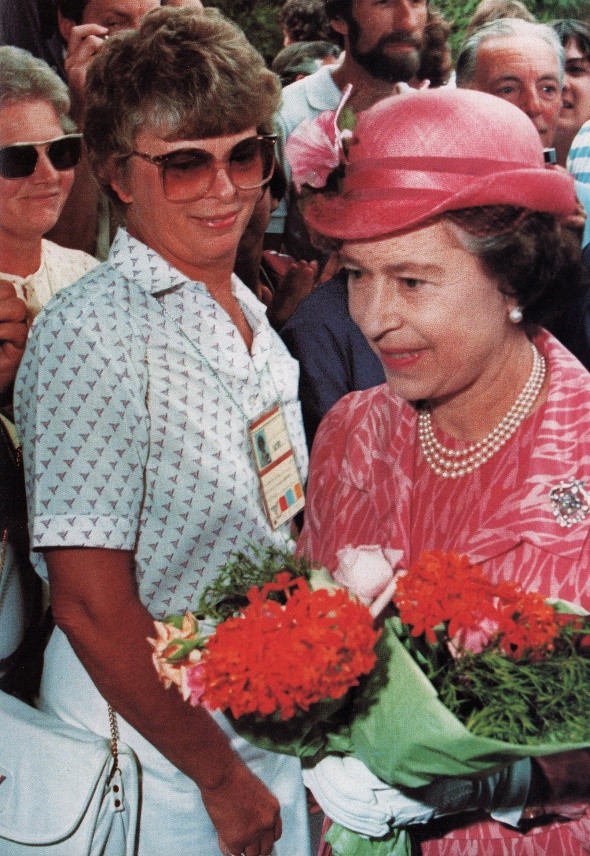 Queen Elizabeth II walks in front of an onlooking crowd wearing a pink outfit and matching hat while holding a bouquet of red flowers.