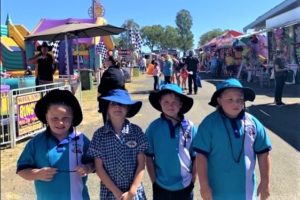 Four children wearing uniforms of the National School for Travelling Show Children. There are rides and games of a sideshow alley behind them. 