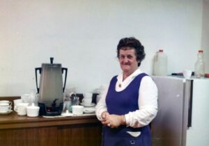 Mrs Taubner is wearing a white shirt and blue smock, standing beside an urn surrounded by crockery. 
