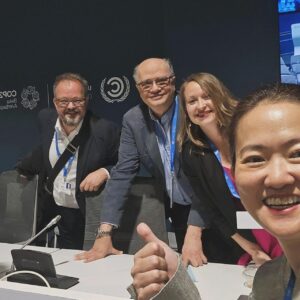 Johanna stands with one woman and two men at a meeting table with logo in the background that reads “COP29 Baku, Azerbaijan”.
