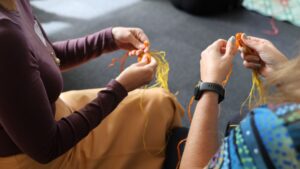 The hands of two people as they weave coloured threads. 